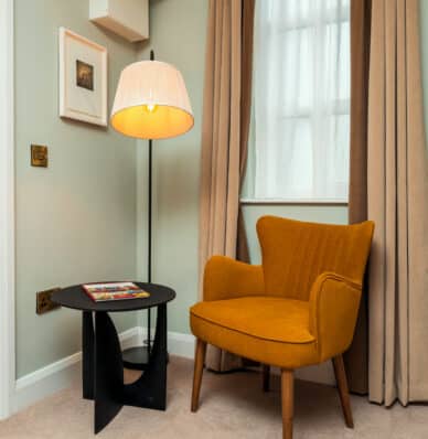 Cosy hotel room corner with a mustard yellow armchair, modern lamp, elegant beige curtains, and a sleek black side table.