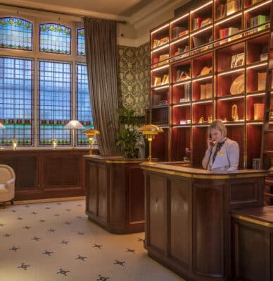 Elegant hotel lobby with wooden desk, staff, stained glass windows, and cosy seating area.