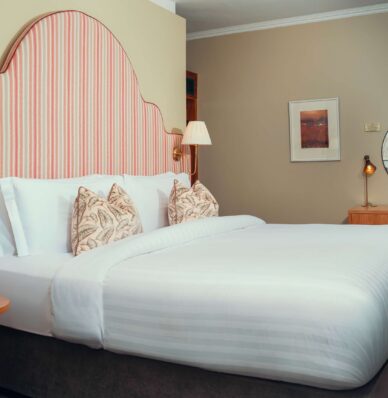 Luxurious hotel bedroom with king-sized bed, stylish decor, and workspace featuring a cosy lamp and art piece.