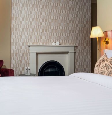 Elegant hotel room with cosy armchair, soft lighting, fireplace, and plush bedding for a relaxing stay.