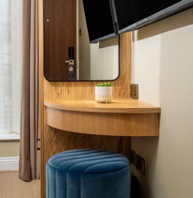 Stylish hotel room with modern wooden desk, mirror, flat-screen TV, plush blue stool, and natural light from large window.