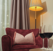 Cosy hotel room corner with plush red armchair, soft lighting, elegant curtains, and two glasses of wine for a relaxing stay.